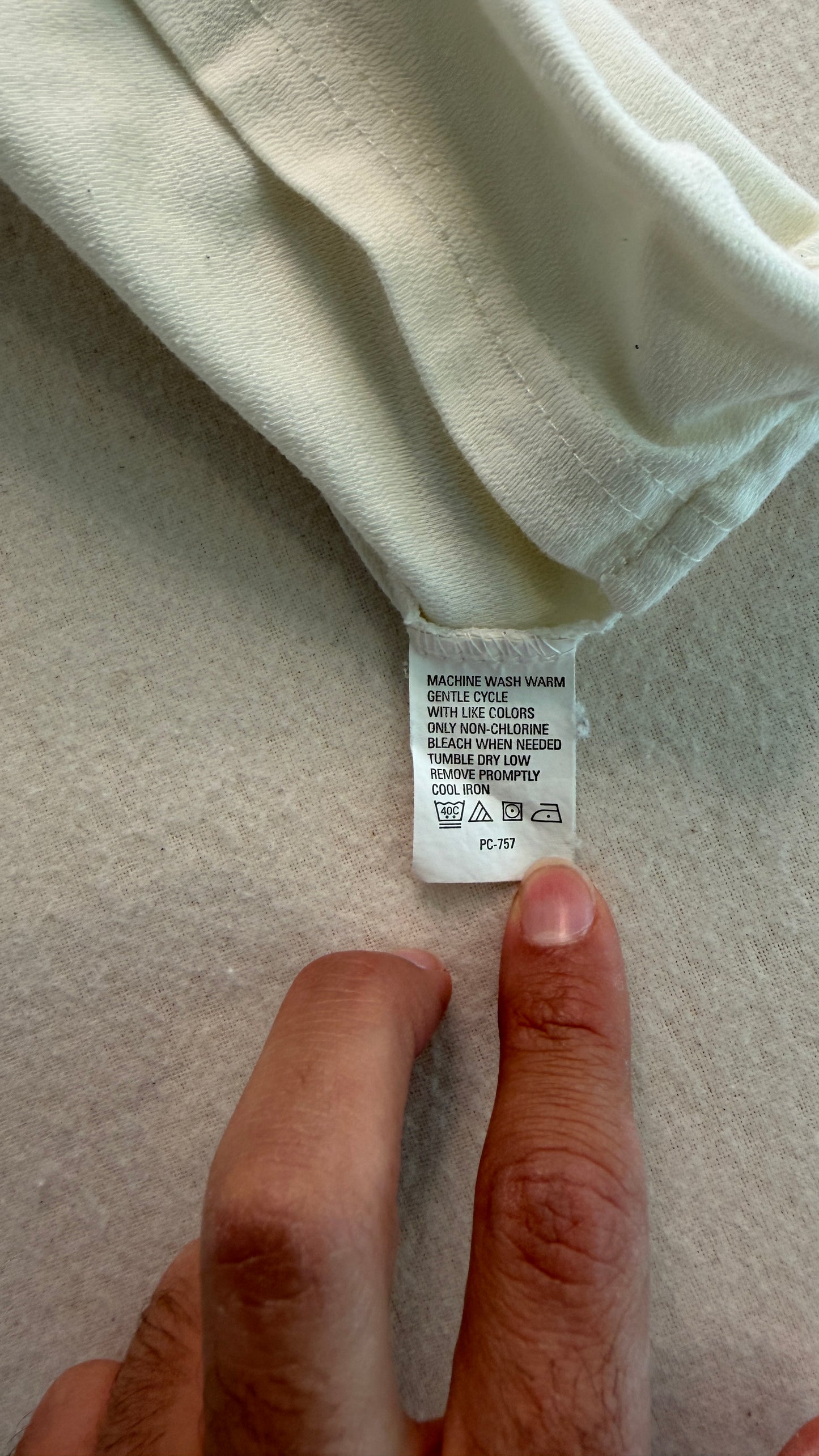 Close-up of a clothing tag on a light-colored fabric with a hand pointing to it.